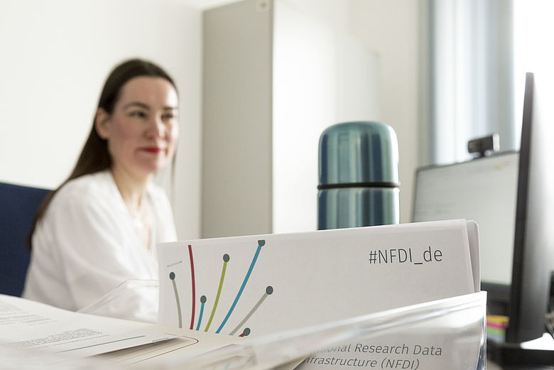 Woman sitting at a desk. On the desk is a display sign with the inscription NFDI_en