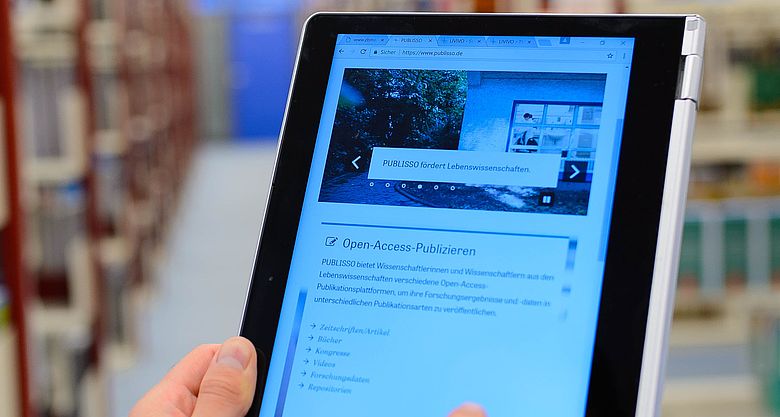 Eine Person, von der nur die Hand zu sehen ist, h&auml;lt ein Tablet, auf dem die Website www.publisso.de zu sehen ist. Im Hintergrund gef&uuml;llte Regale in einer Bibliohek.